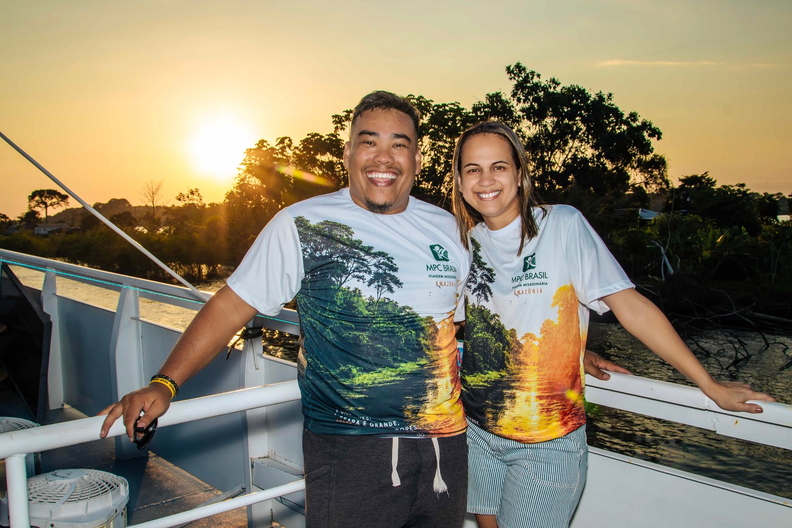 Marcos e Aline sorrindo, com camisetas da viagem missionária MPC Brasil Amazônia, ao entardecer